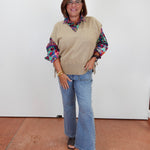 A woman modeling a beige knit sweater vest over a colorful patterned long-sleeve blouse, paired with light blue wide-leg jeans and black sandals, standing against a plain white wall.