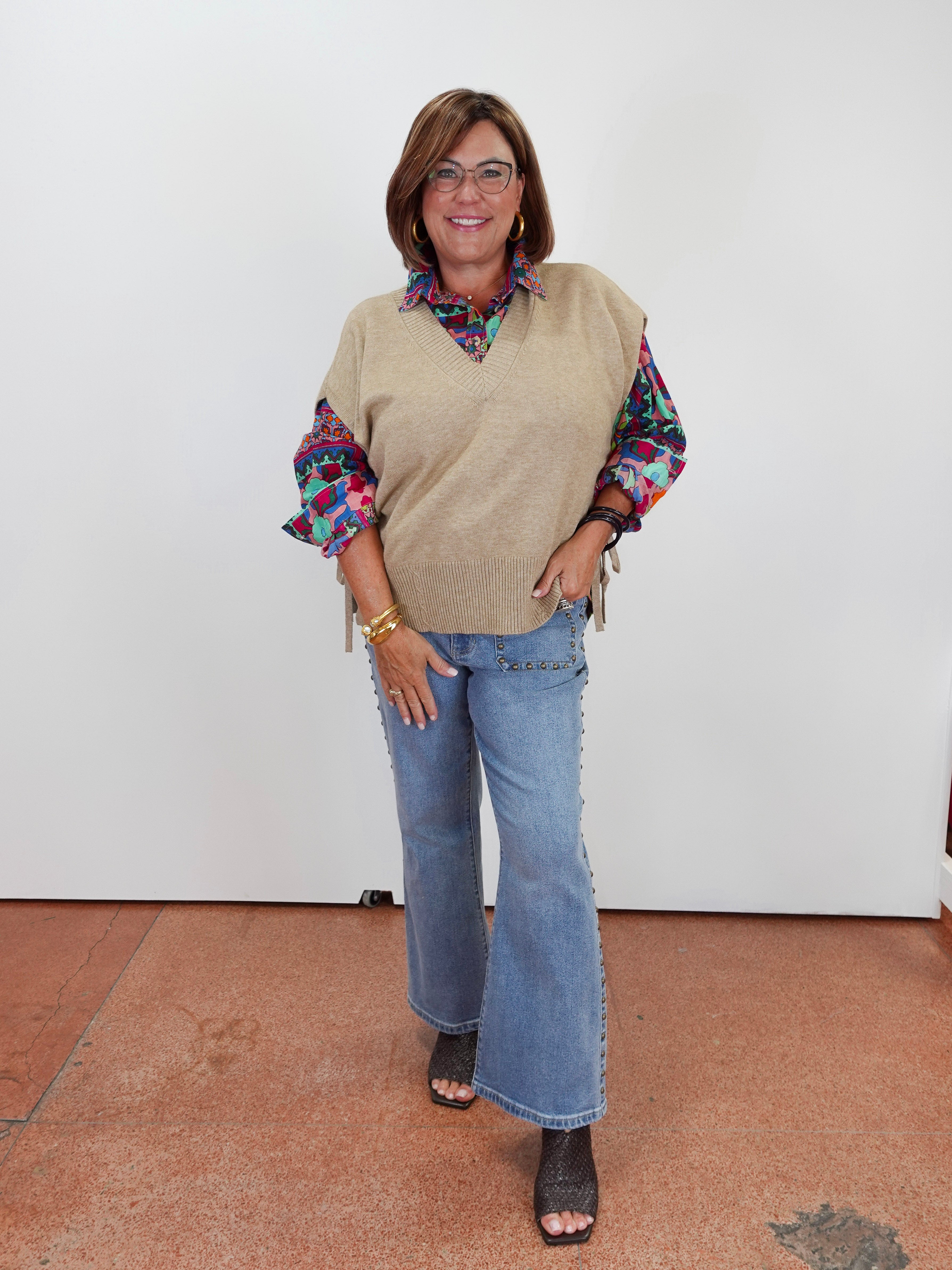 A woman modeling a beige knit sweater vest over a colorful patterned long-sleeve blouse, paired with light blue wide-leg jeans and black sandals, standing against a plain white wall.