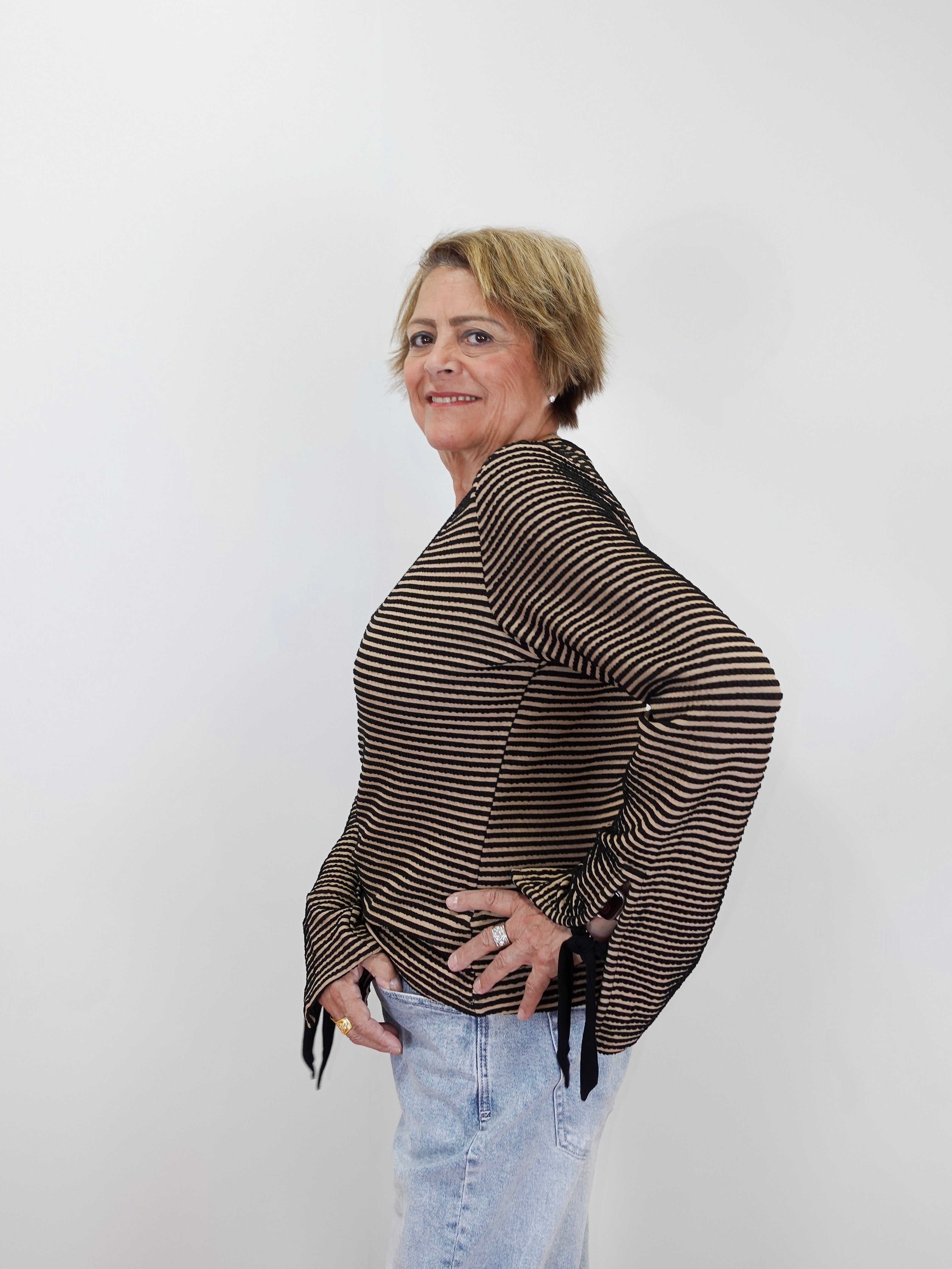 A woman with short blonde hair models a brown and black striped long-sleeve top with tie details at the cuffs, paired with light blue jeans, against a plain white background.