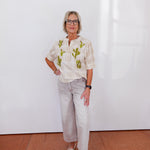 Woman wearing a patterned blouse and light pants against a white wall.