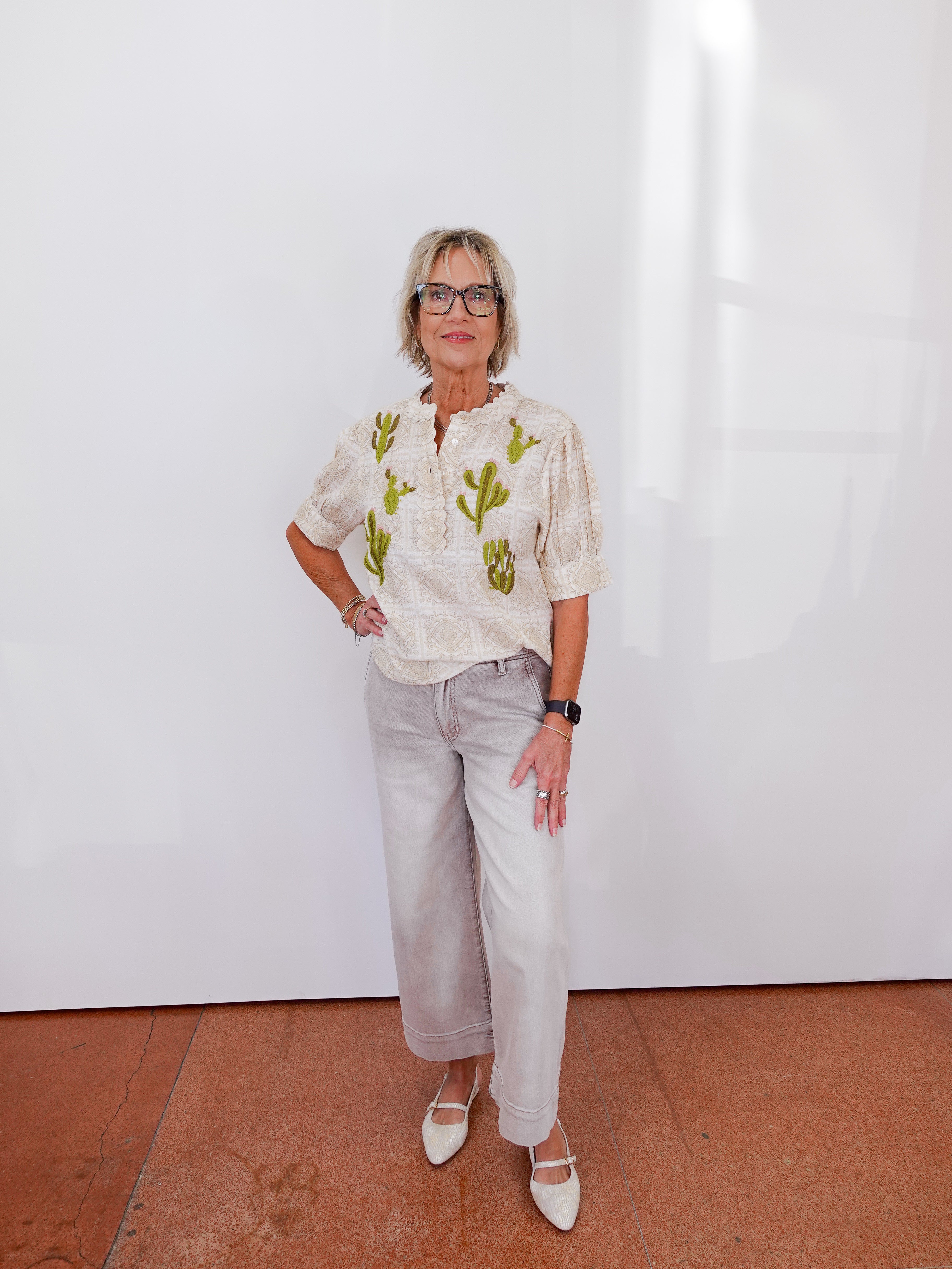 Woman wearing a patterned blouse and light pants against a white wall.