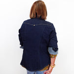 A woman viewed from behind wearing a dark indigo denim shirt jacket over a patterned top, paired with light blue jeans and holding a red handbag against a plain white background.