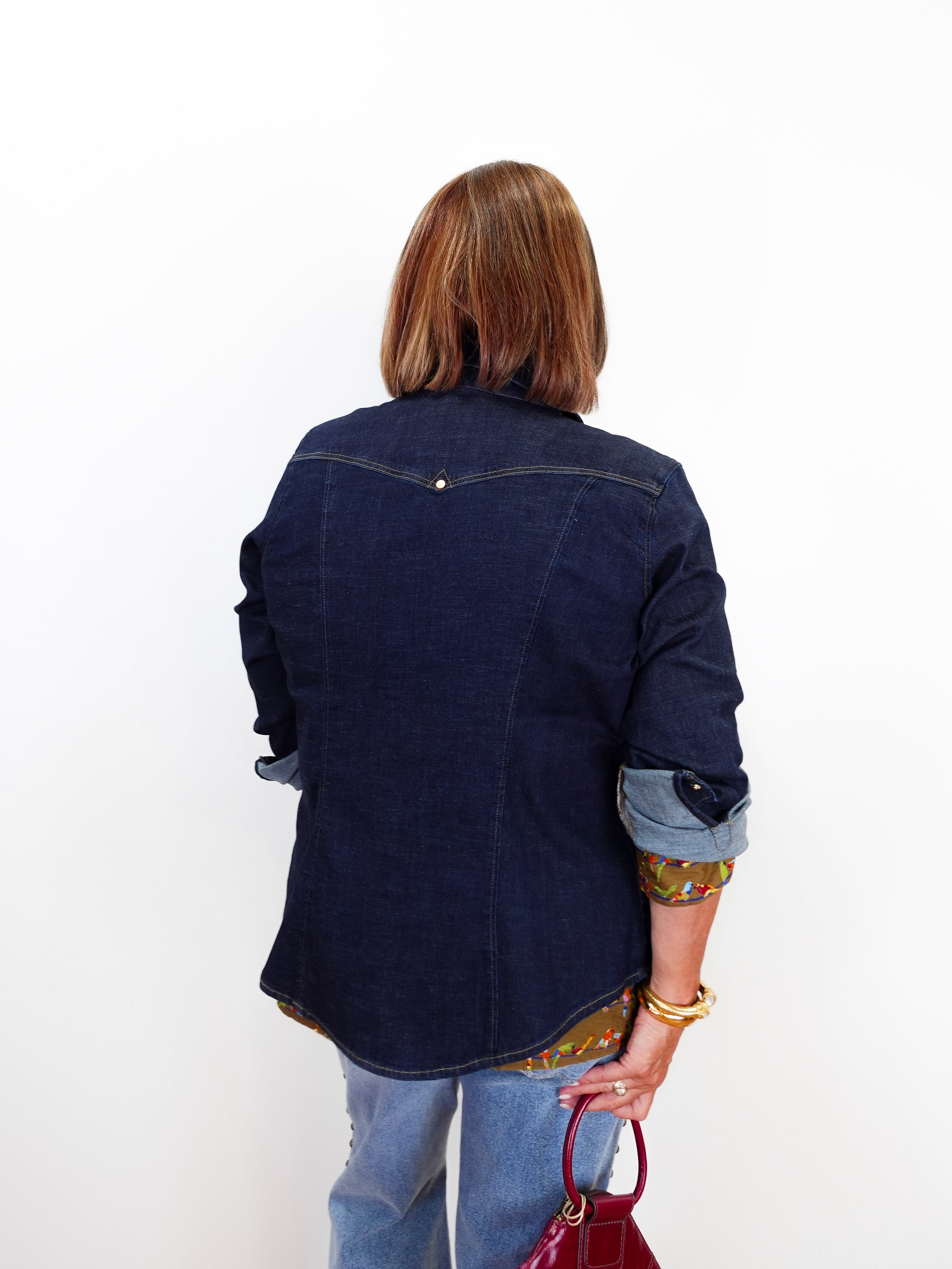 A woman viewed from behind wearing a dark indigo denim shirt jacket over a patterned top, paired with light blue jeans and holding a red handbag against a plain white background.