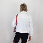 Person wearing a white jacket and black shorts with a colorful bag over one shoulder against a plain background