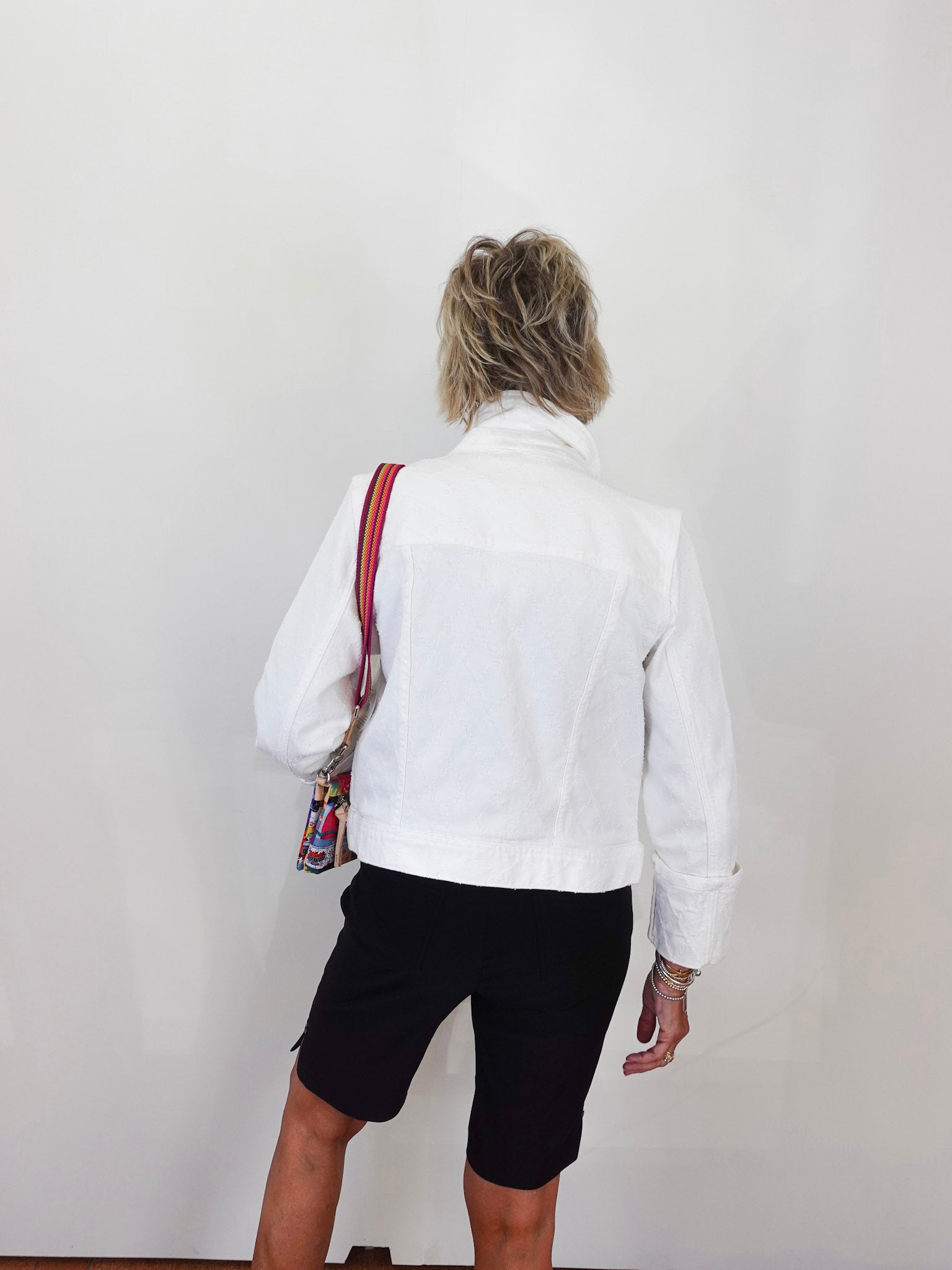 Person wearing a white jacket and black shorts with a colorful bag over one shoulder against a plain background