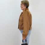 A woman in profile wearing a brown suede jacket with lace trim, light blue jeans, and holding a black handbag, standing against a plain white wall.