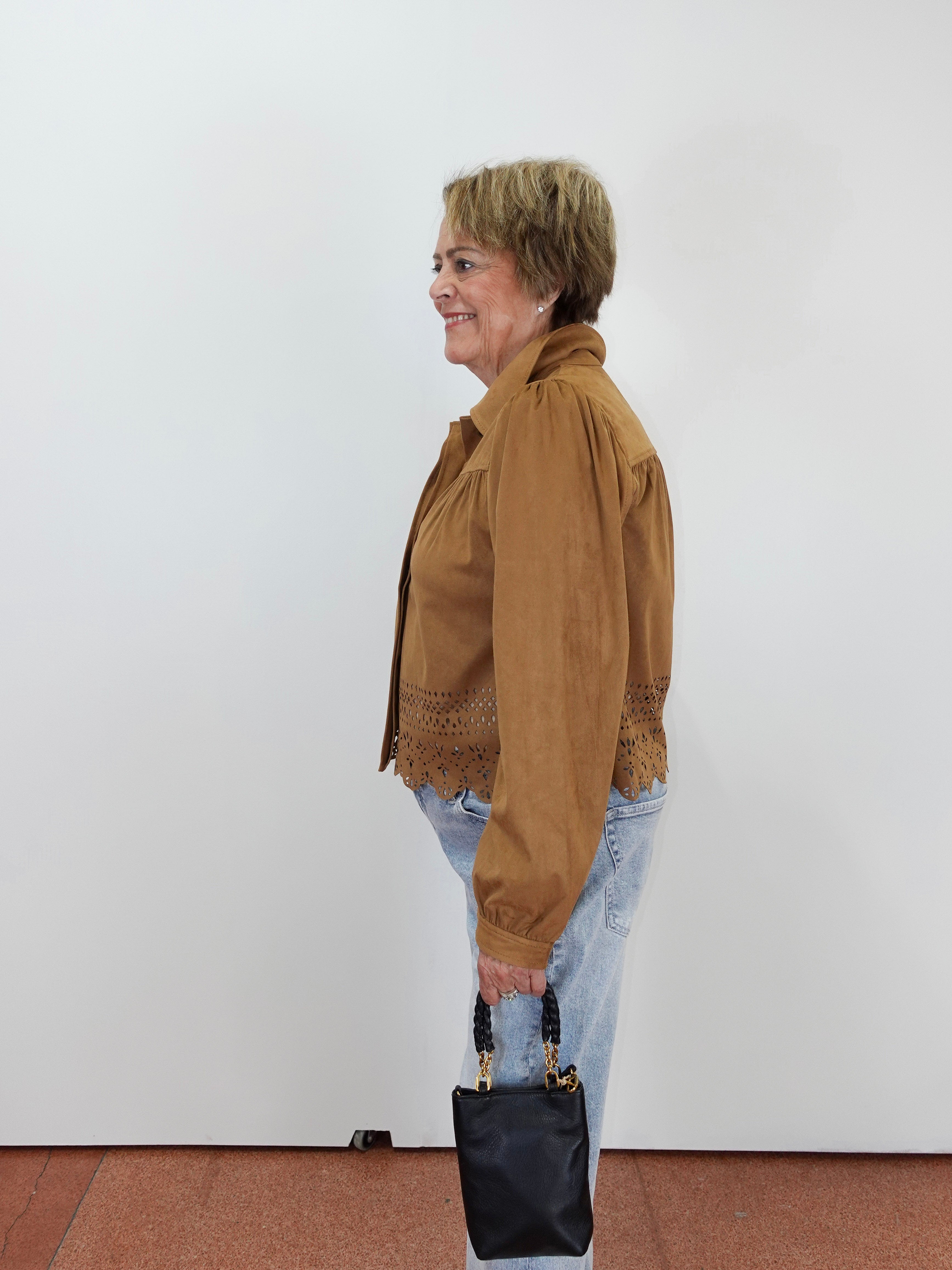 A woman in profile wearing a brown suede jacket with lace trim, light blue jeans, and holding a black handbag, standing against a plain white wall.