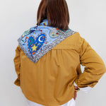A woman viewed from behind wearing a mustard yellow jacket over a white shirt, with a colorful patterned scarf draped around her neck. The scarf features a blue heart, crescent moon, star, and decorative border.
