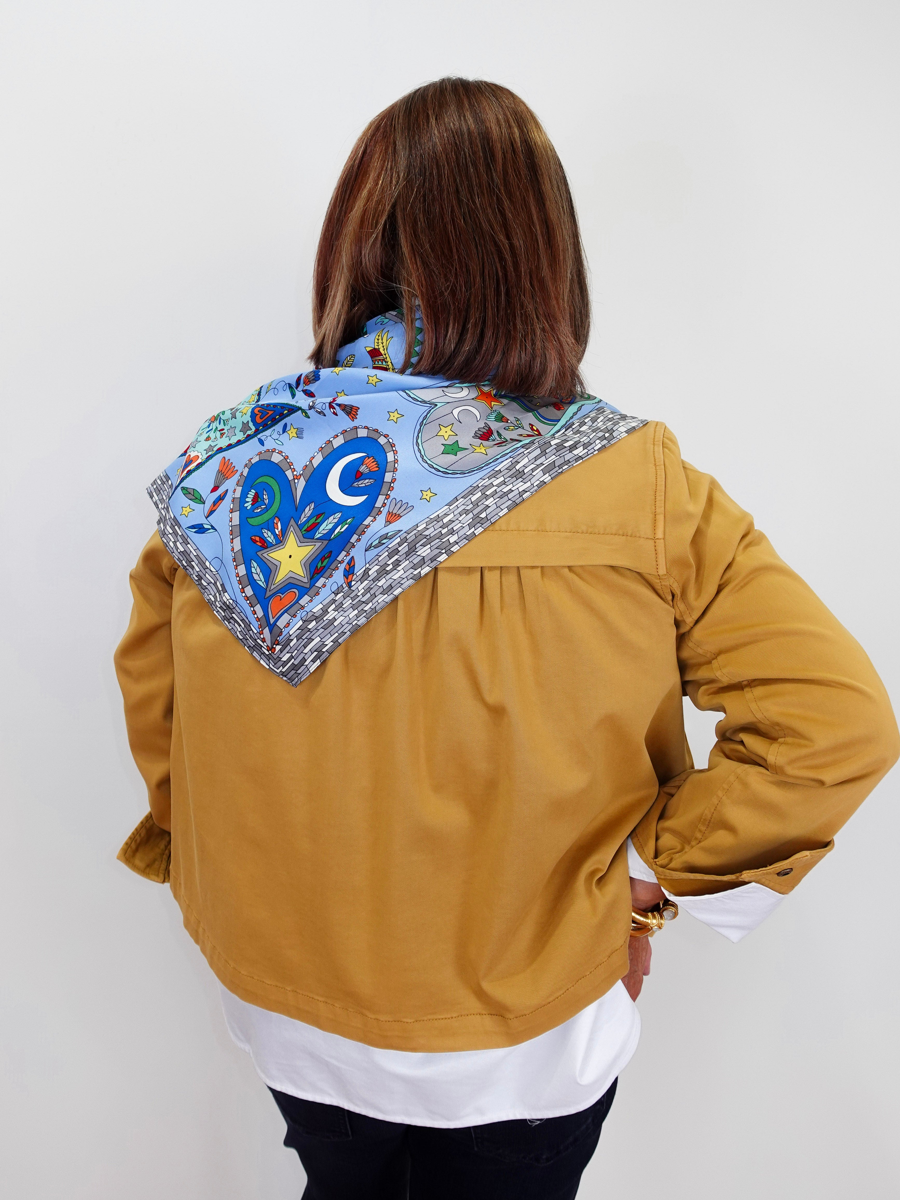 A woman viewed from behind wearing a mustard yellow jacket over a white shirt, with a colorful patterned scarf draped around her neck. The scarf features a blue heart, crescent moon, star, and decorative border.