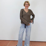 A woman stands against a plain white wall wearing a dark olive-brown and beige horizontally striped long-sleeve top with light-wash wide-leg jeans and black sandals.