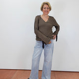 A woman stands against a plain white wall wearing a dark olive-brown and beige horizontally striped long-sleeve top with light-wash wide-leg jeans and black sandals.