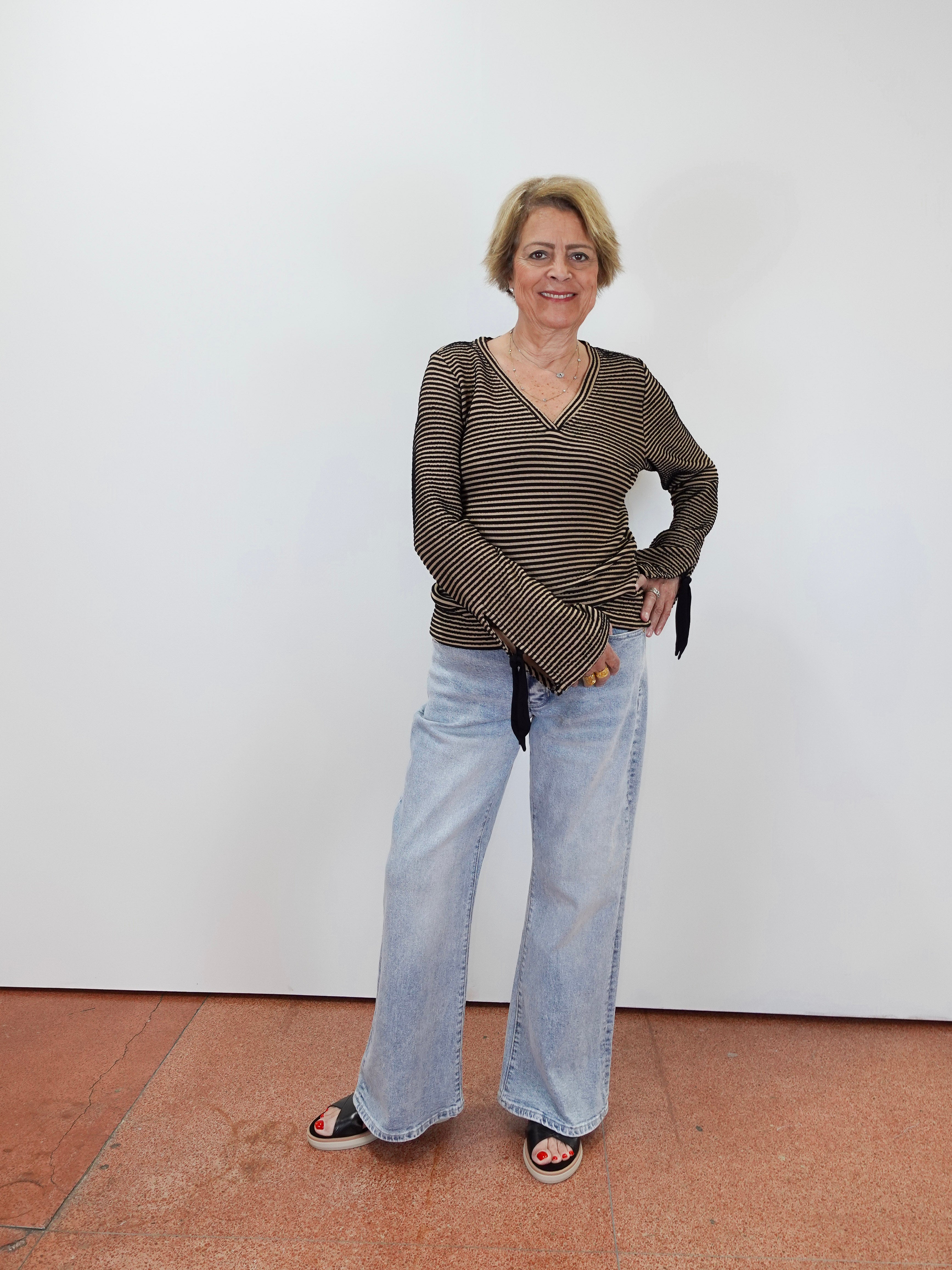 A woman stands against a plain white wall wearing a dark olive-brown and beige horizontally striped long-sleeve top with light-wash wide-leg jeans and black sandals.
