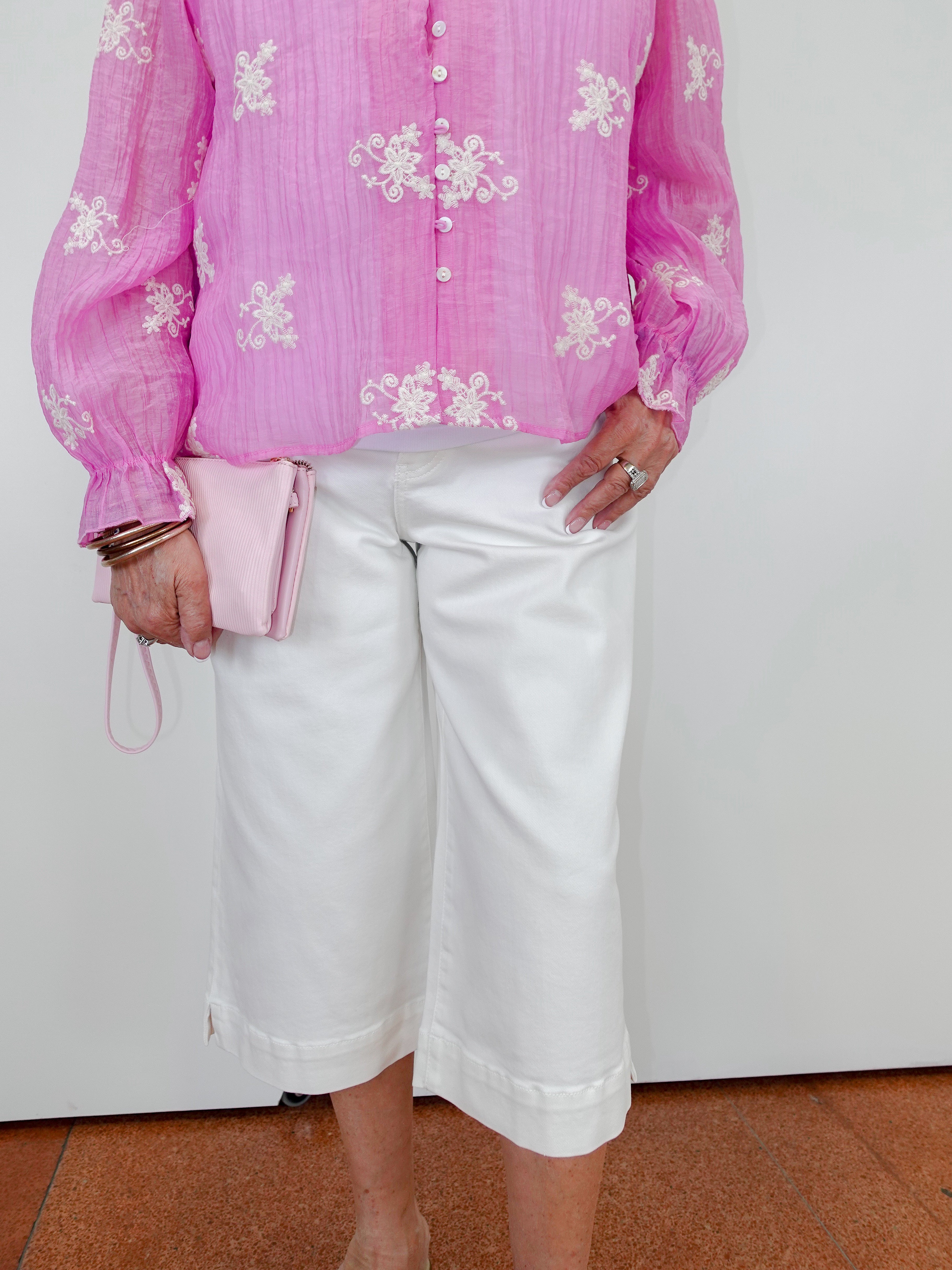 Person wearing a pink blouse with white floral patterns and white pants against a plain background