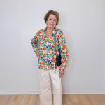 Woman wearing a colorful patterned jacket and white pants against a plain white wall.
