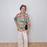 Woman wearing a colorful patterned jacket and white pants against a plain white wall.