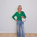 Woman wearing a green shirt and blue jeans standing against a white wall.