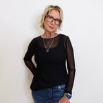 Woman wearing a black long-sleeve top with sheer sleeves and blue jeans against a white background