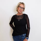 Woman wearing a black long-sleeve top with sheer sleeves and blue jeans against a white background