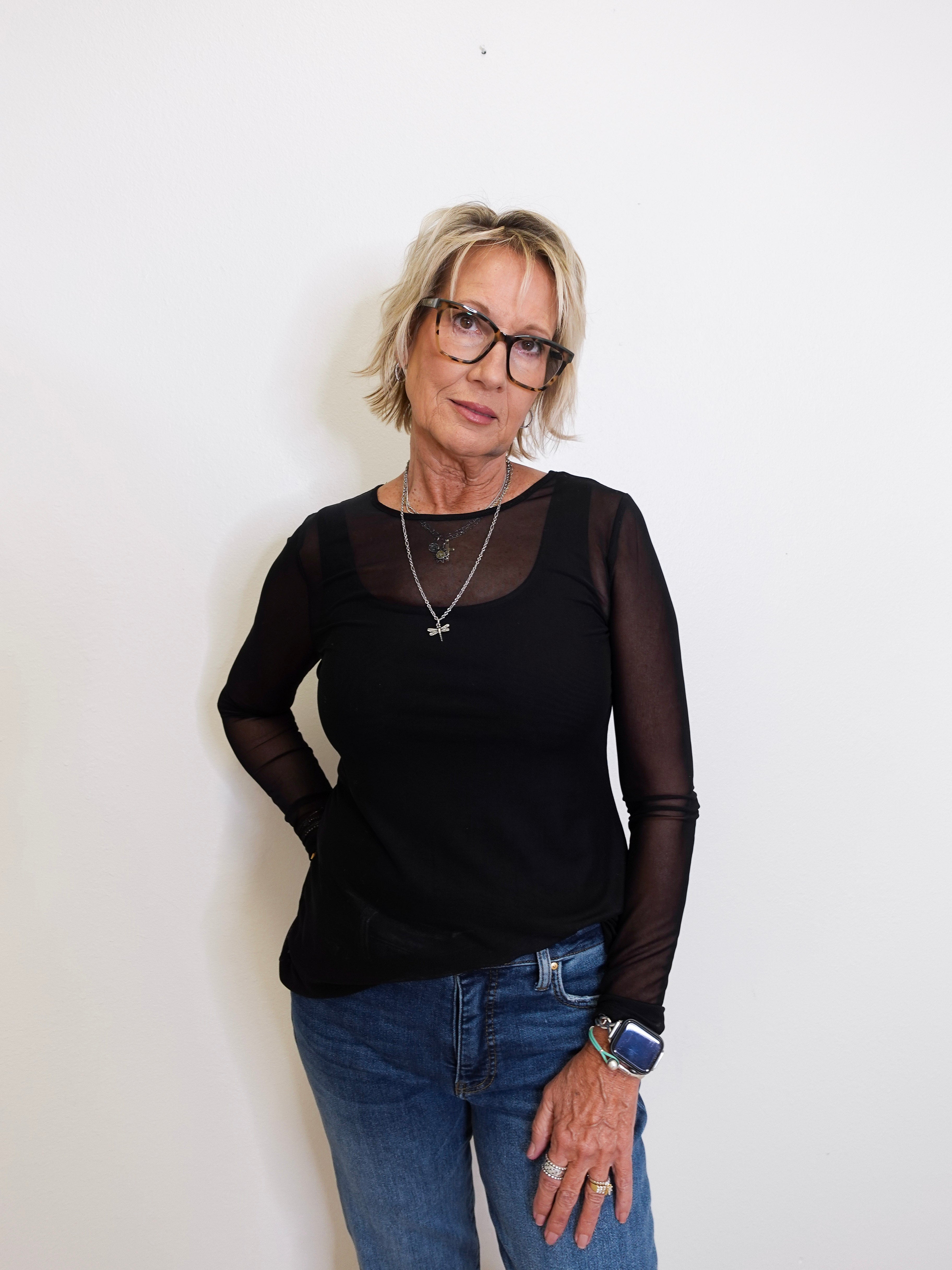 Woman wearing a black long-sleeve top with sheer sleeves and blue jeans against a white background