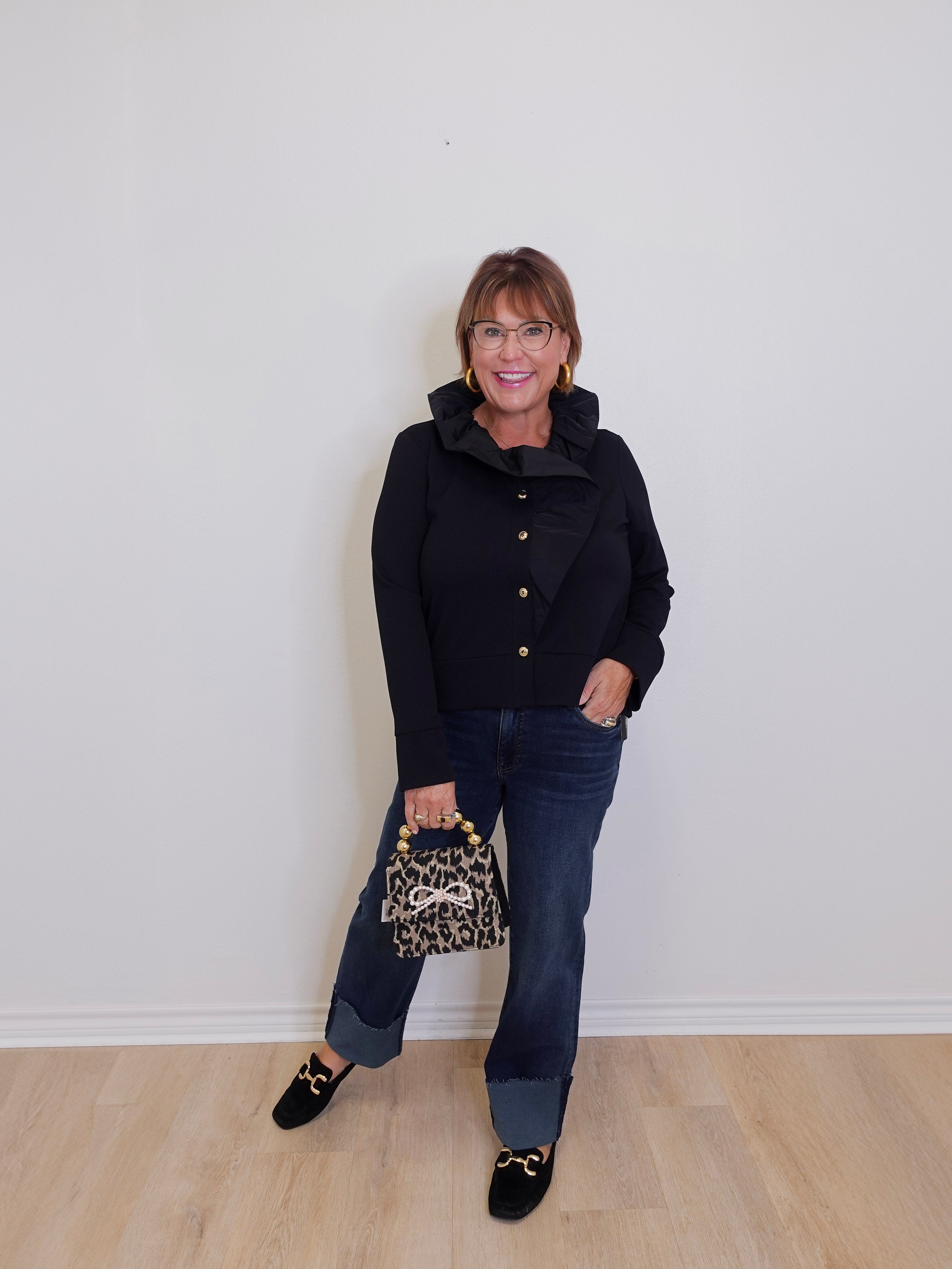 Woman wearing a black coat and jeans, holding a leopard print handbag against a white wall.