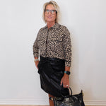 Person wearing a leopard print shirt and black leather skirt holding a black handbag against a white wall.