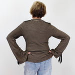 A person viewed from behind wearing a brown and beige horizontally striped long-sleeve top with flared cuffs and light blue jeans, standing against a plain white wall.
