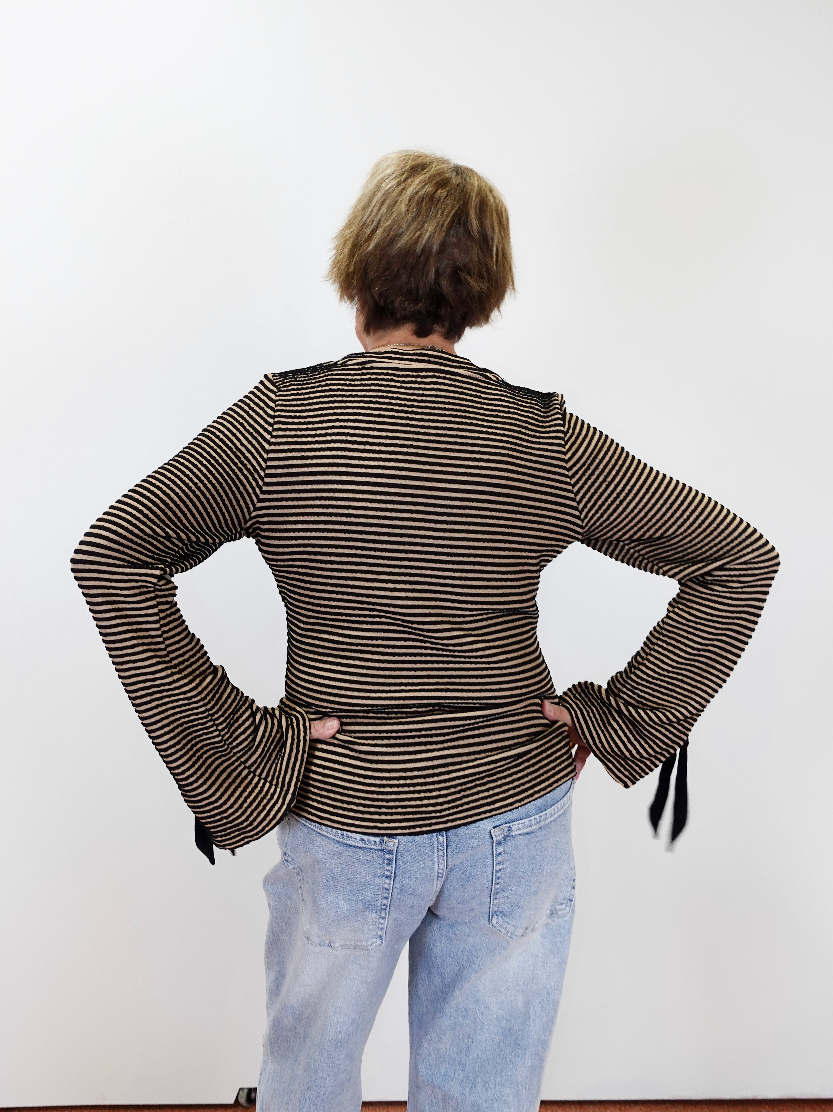 A person viewed from behind wearing a brown and beige horizontally striped long-sleeve top with flared cuffs and light blue jeans, standing against a plain white wall.
