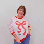 Woman wearing a white sweater with a red bow design and blue jeans against a plain background