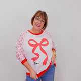 Woman wearing a white sweater with a red bow design and blue jeans against a plain background