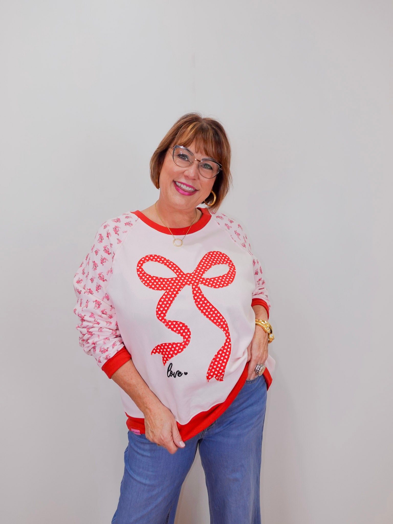 Woman wearing a white sweater with a red bow design and blue jeans against a plain background