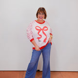 Woman wearing a white sweater with red accents and blue pants against a plain background