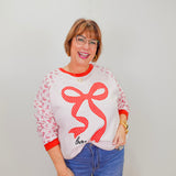 Woman wearing a white sweatshirt with a red bow design and blue jeans against a plain background