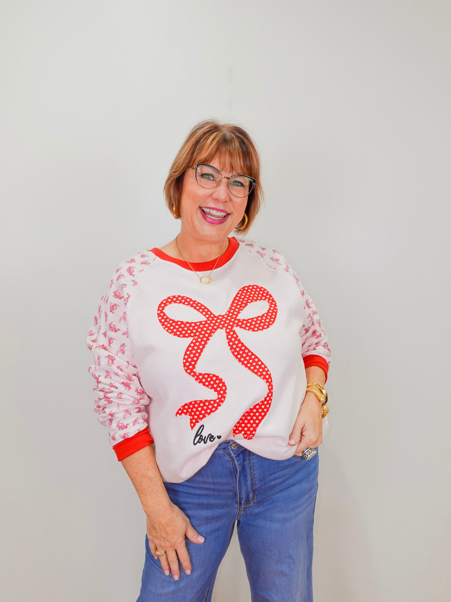 Woman wearing a white sweatshirt with a red bow design and blue jeans against a plain background