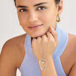 Woman wearing a silver heart-shaped necklace and gold earrings against a neutral background