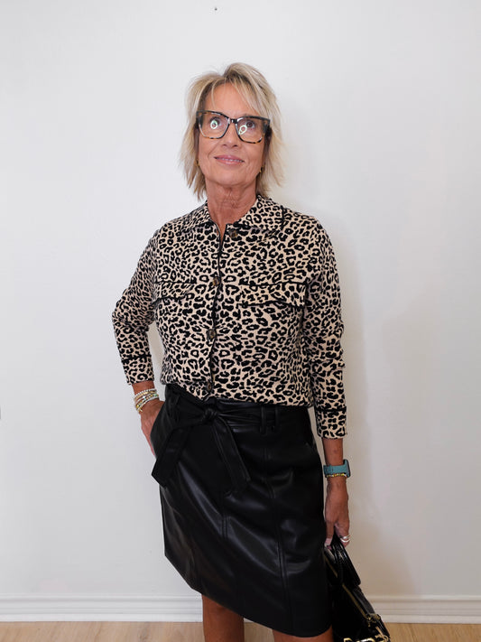 Woman wearing a leopard print shirt and black skirt against a white wall.