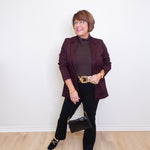 Woman wearing a dark purple blazer and black pants against a white wall.