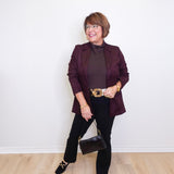 Woman wearing a dark purple blazer and black pants against a white wall.