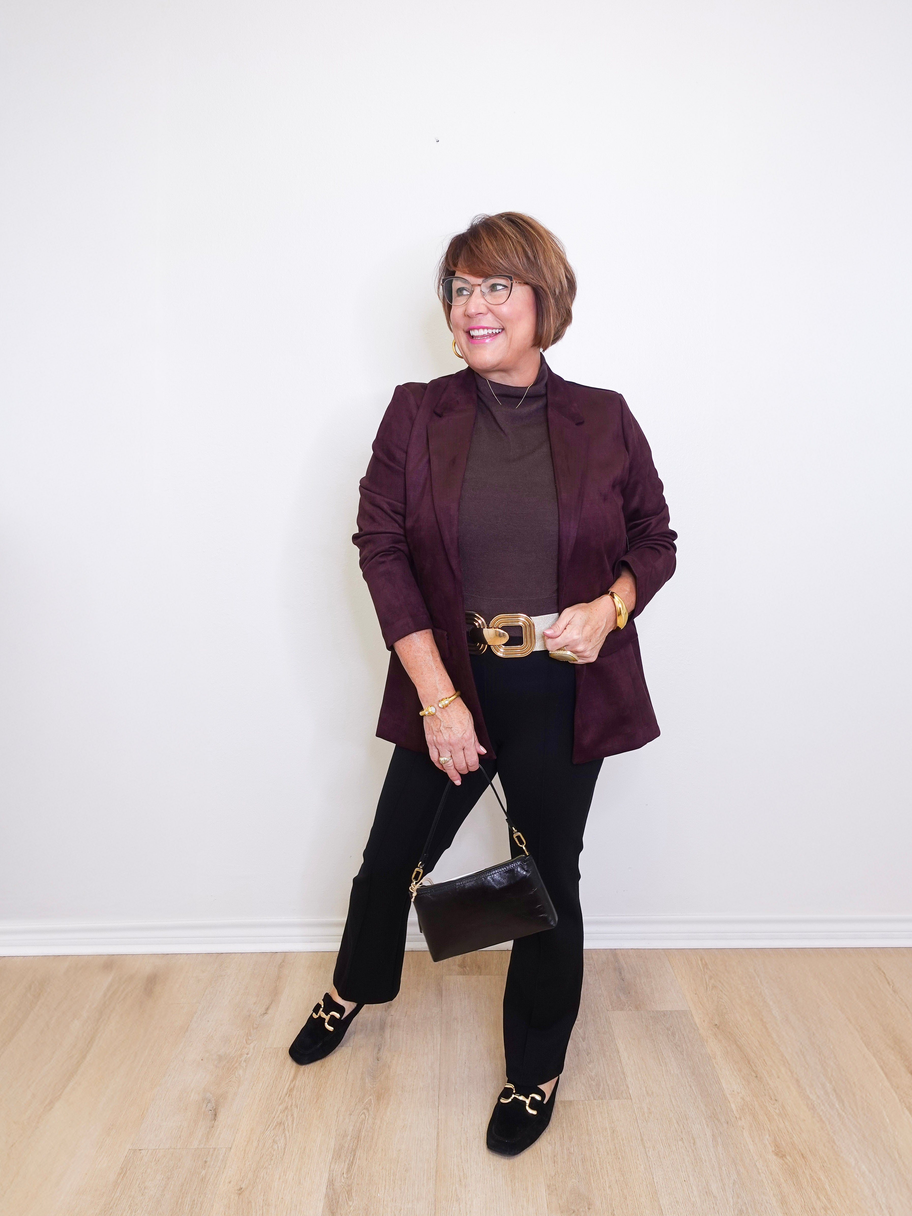 Woman wearing a dark purple blazer and black pants against a white wall.