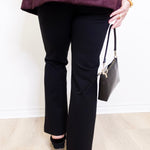 Person wearing black pants and a purple top, holding a black handbag against a white background