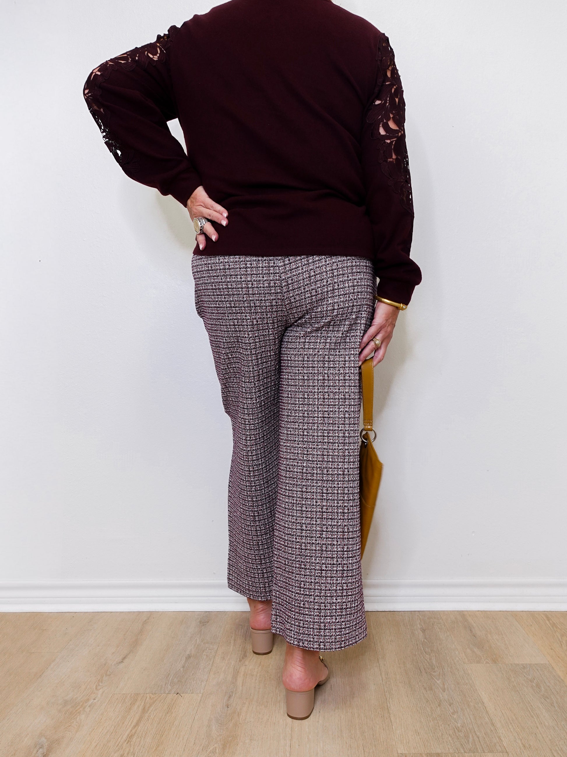 Person wearing a dark brown sweater with lace sleeves and patterned pants against a white wall.