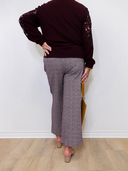 Person wearing a dark brown sweater with lace sleeves and patterned pants against a white wall.