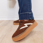 Brown sneakers with white accents worn with blue jeans on a light wooden floor.