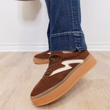 Brown sneakers with white accents worn with blue jeans on a light wooden floor.