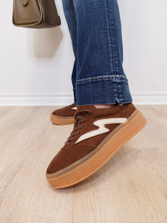 Brown sneakers with white accents worn with blue jeans on a light wooden floor.