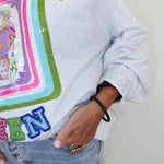 A close-up of a person wearing a light gray heather sweatshirt with a large colorful sequined graphic and the word 'MAJJOVC' across the chest, paired with light blue jeans.