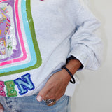 A close-up of a person wearing a light gray heather sweatshirt with a large colorful sequined graphic and the word 'MAJJOVC' across the chest, paired with light blue jeans.