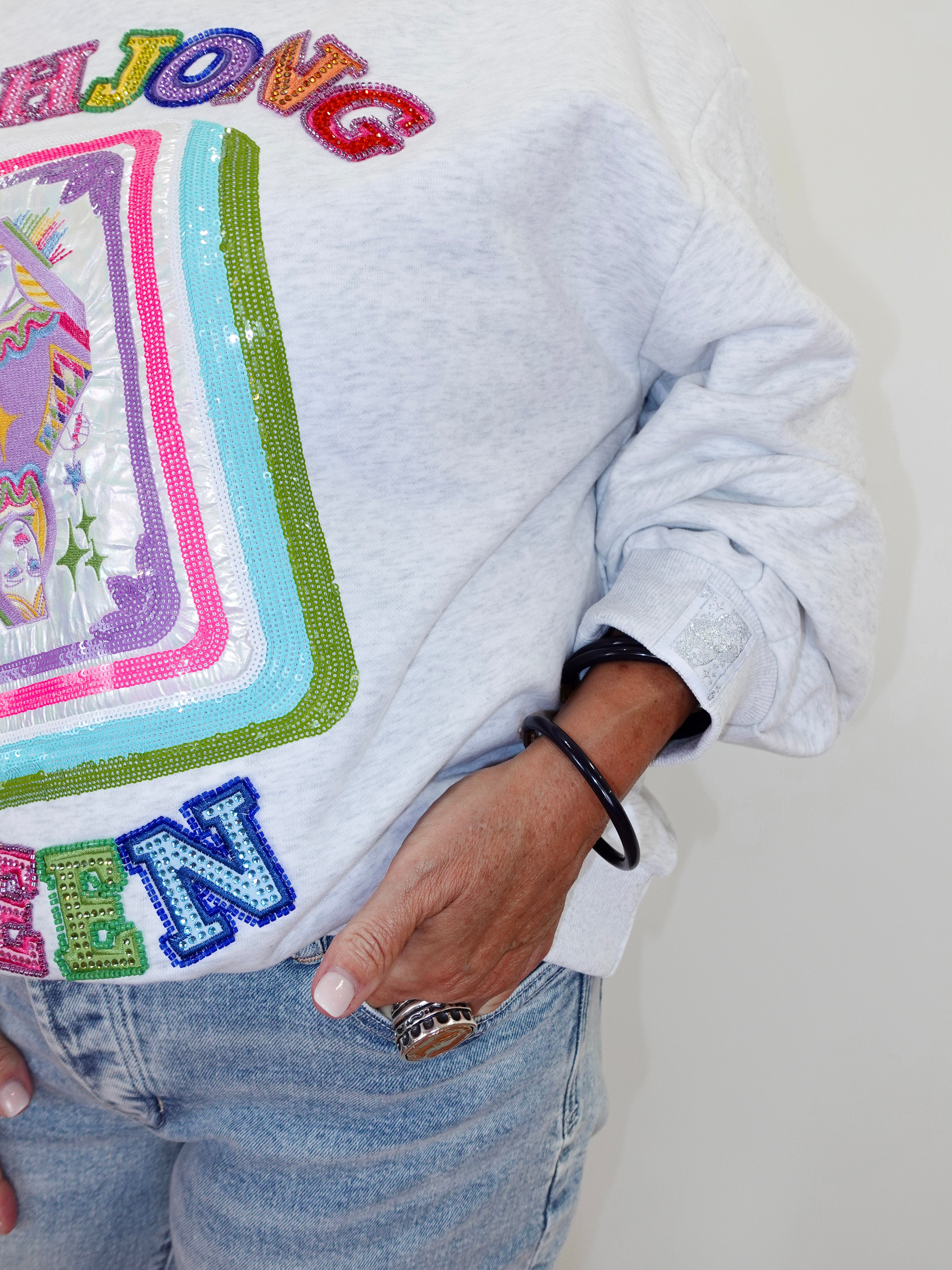 A close-up of a person wearing a light gray heather sweatshirt with a large colorful sequined graphic and the word 'MAJJOVC' across the chest, paired with light blue jeans.