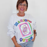 A woman wearing a white long-sleeve sweatshirt with a colorful 'Mahjong Queen' graphic on the front, paired with light-wash jeans, standing against a plain white wall.