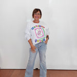 A woman stands against a white wall wearing a white long-sleeve graphic sweatshirt with a colorful retro design, light-wash flare jeans, and beige sandals.