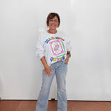 A woman stands against a white wall wearing a white long-sleeve graphic sweatshirt with a colorful retro design, light-wash flare jeans, and beige sandals.